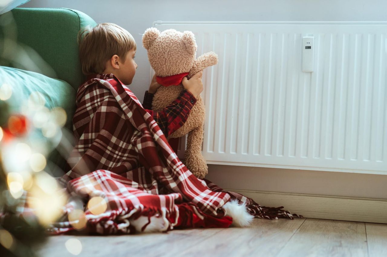 Ребенок в квартире с игрушкой мишкой сидит на полу и трогает батарею, отопление
