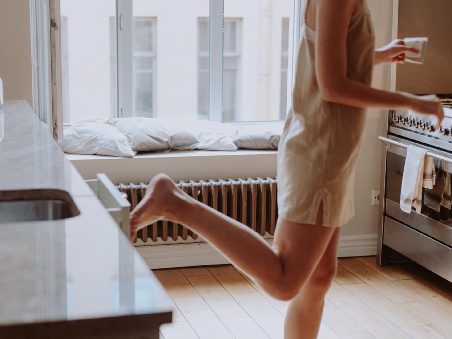 Девушка на кухне в халате закрывает ногой ящик, батарея на заднем плане, окно, солнце