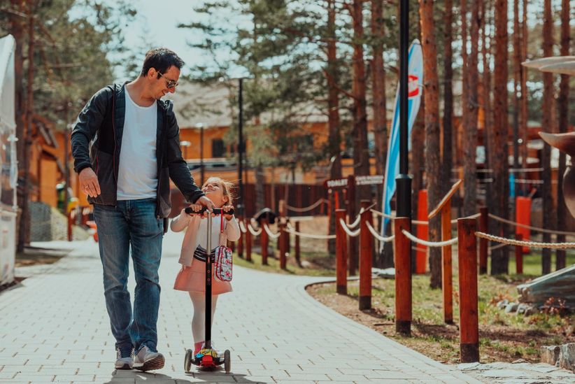 Отец с дочкой гуляют по парку, девочка на самокате